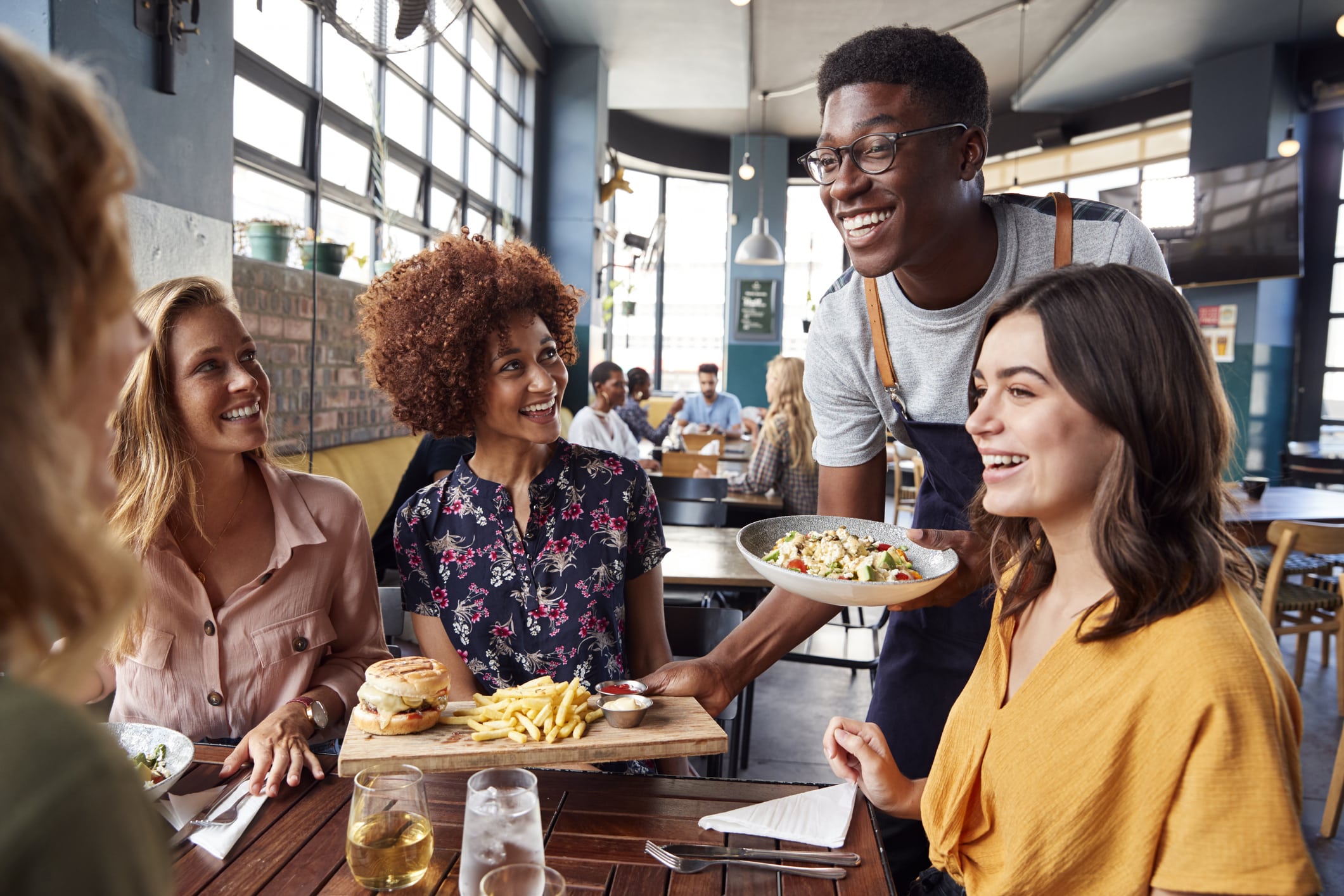 Un serveur souriant sert un repas à un groupe de quatre amis ravis, assis à une table de restaurant, qui discutent et savourent leur repas dans un cadre lumineux et moderne.