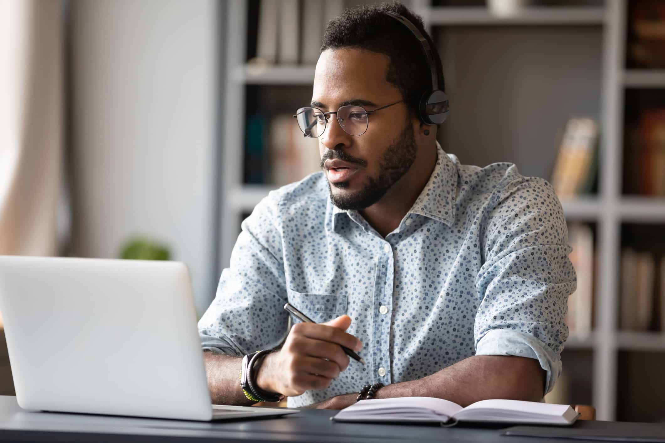 Un homme portant des lunettes et des écouteurs est assis à un bureau, les yeux rivés sur l'écran d'un ordinateur portable. Il tient un stylo à la main et a un cahier ouvert devant lui ; il semble concentré et absorbé par une réunion virtuelle ou un cours en ligne.