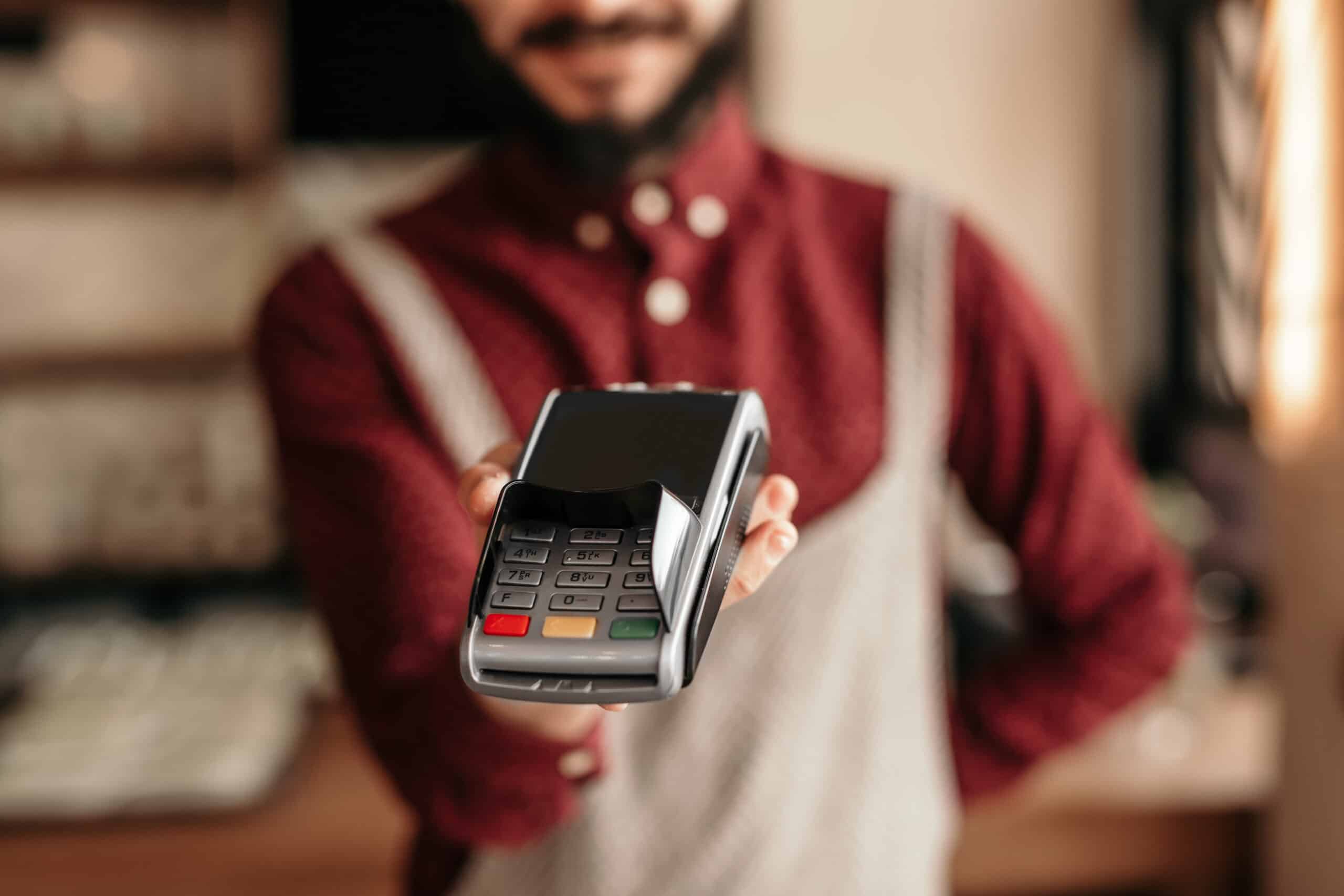Une personne vêtue d'une chemise rouge et d'un tablier tient un terminal de paiement par carte face à la caméra, prête à effectuer un paiement sans contact. Le terminal est net, tandis que la personne est floue à l'arrière-plan.