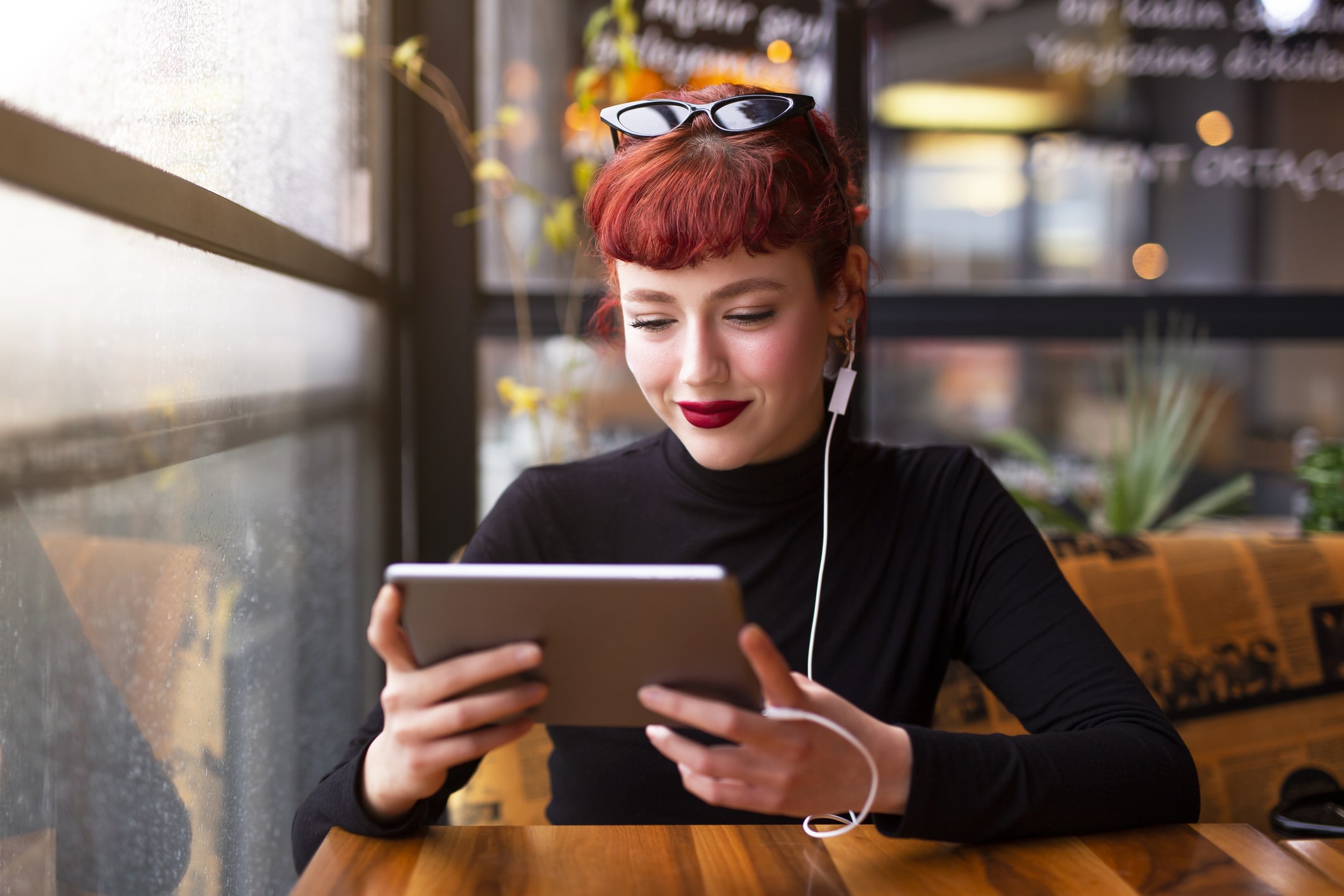 Une femme aux cheveux roux, des lunettes de soleil posées sur la tête, est assise à la table d'un café ; elle sourit en regardant sa tablette, des écouteurs aux oreilles.