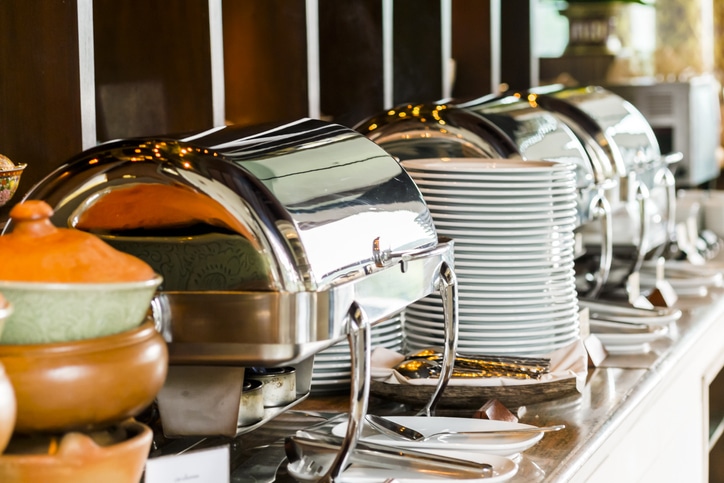 Un buffet composé de réchauds couverts, de piles d'assiettes blanches, d'ustensiles de service et de bols en céramique disposés sur un plan de travail, prêt à accueillir les convives qui viendront se servir eux-mêmes.