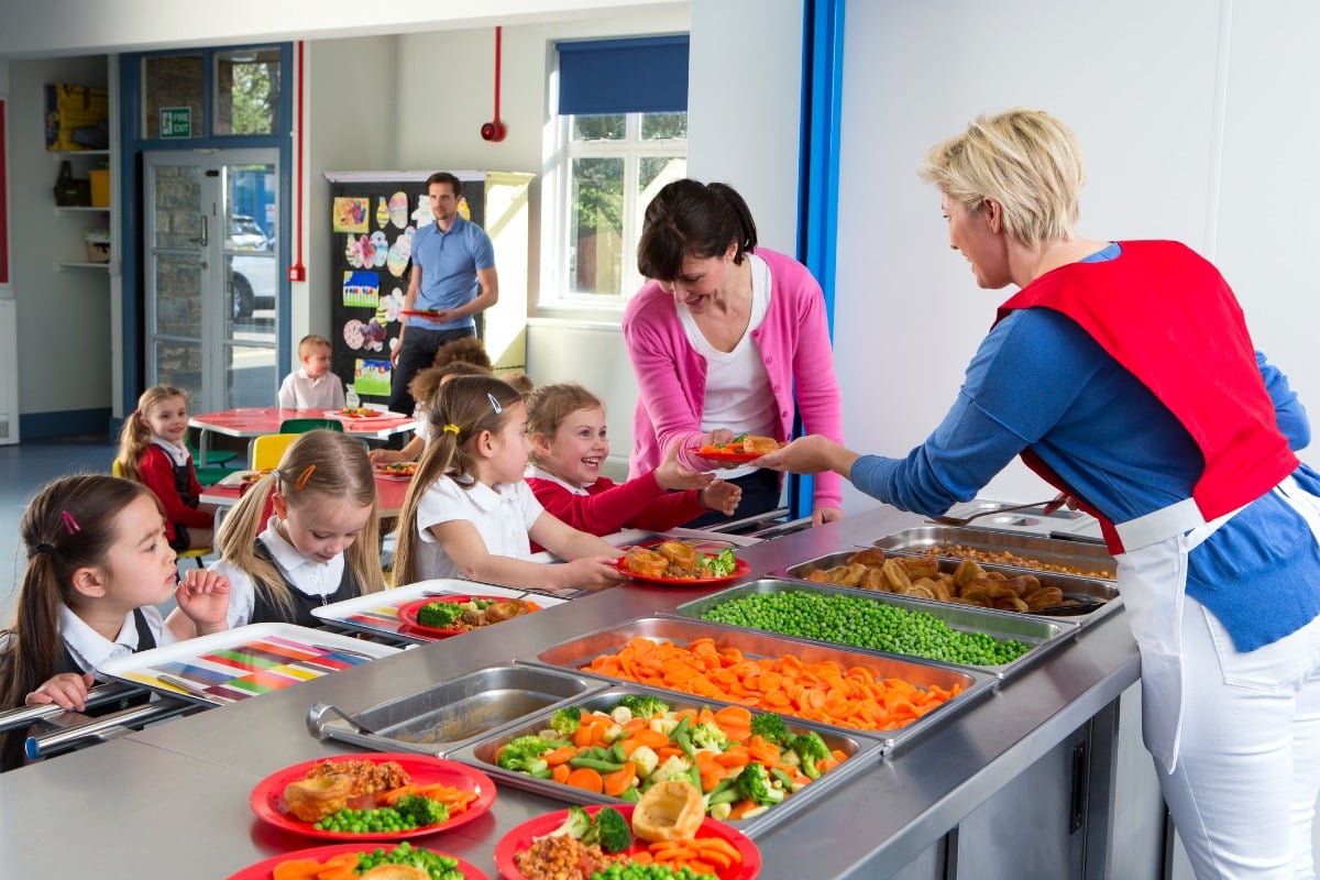 Dans la file d'attente de la cantine scolaire, des enfants reçoivent leur repas des mains d'un serveur. Les assiettes sont remplies de petits pois, de carottes et d'autres aliments. Un accompagnateur aide à servir les repas, et les enfants s'assoient à table pour déjeuner.