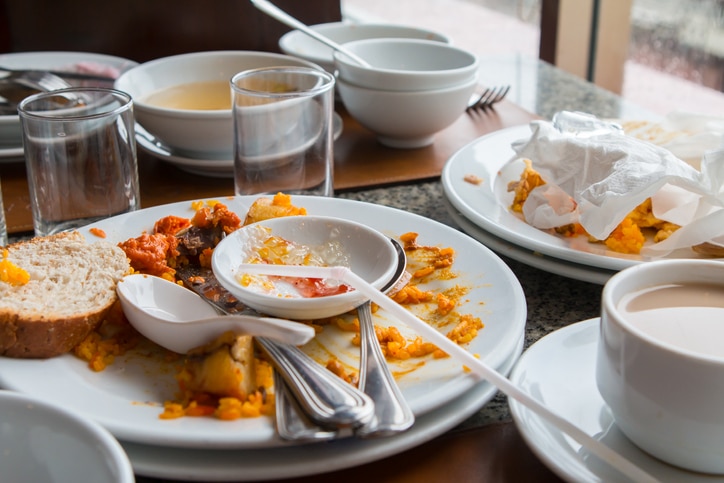 Plusieurs assiettes sales, bols, couverts, verres vides et une tasse de café sont posés sur une table après le repas. Certaines assiettes contiennent des restes de nourriture et des serviettes, ce qui indique que le repas est terminé.