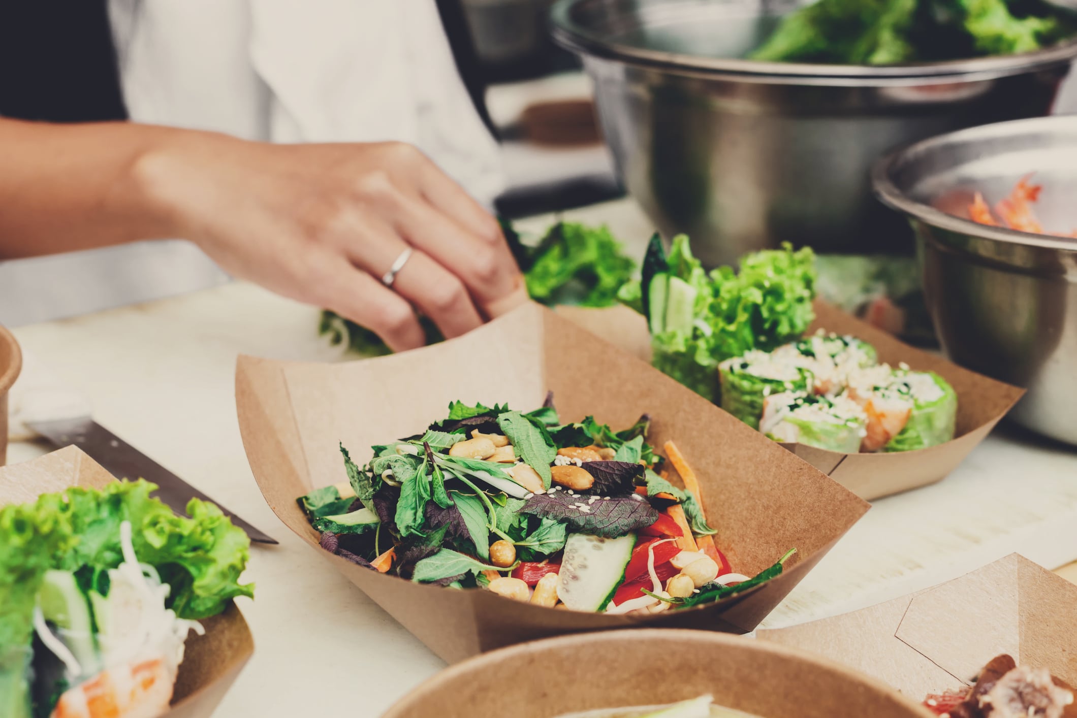 Une main prépare une salade fraîche dans un plateau en papier kraft, entourée d'autres plateaux remplis de légumes verts à feuilles et de légumes, sur un plan de travail de cuisine.