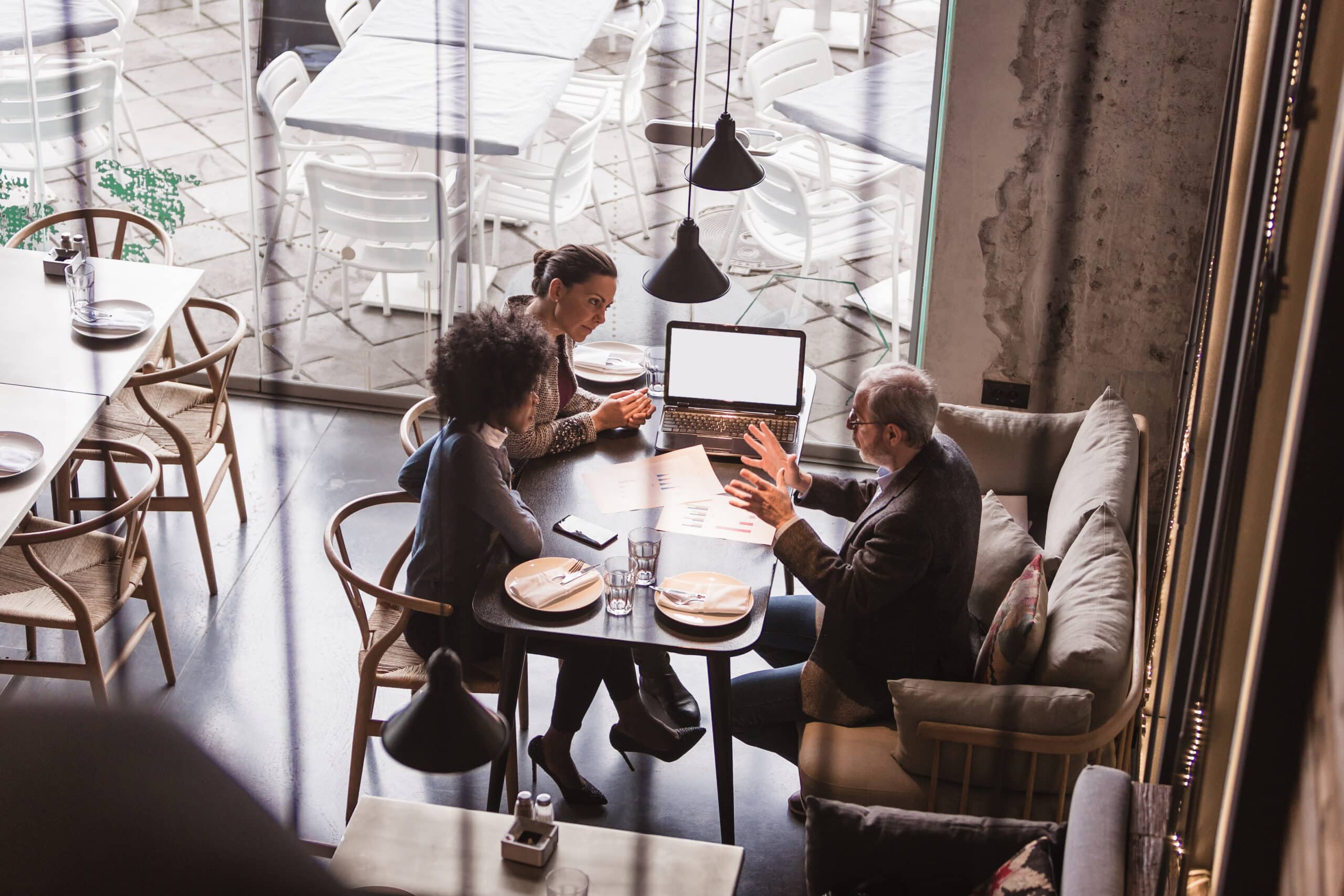 Trois personnes sont assises à une table de restaurant, un ordinateur portable et des documents devant elles, en pleine discussion. Le décor est moderne : la lumière naturelle pénètre par de grandes baies vitrées et on aperçoit des tables vides à l'extérieur.