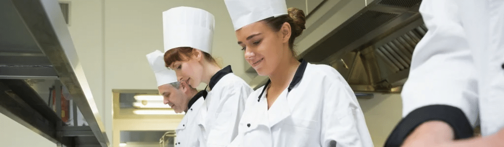 Trois chefs, vêtus d'uniformes blancs et coiffés de toques, travaillent dans une cuisine professionnelle, concentrés sur la préparation des plats derrière un comptoir baigné d'une lumière vive.