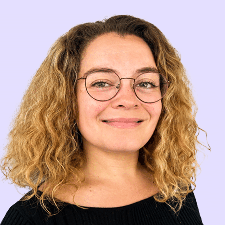Une femme aux cheveux blonds bouclés et portant des lunettes sourit à la caméra. Elle porte un haut noir et pose devant un fond violet clair.