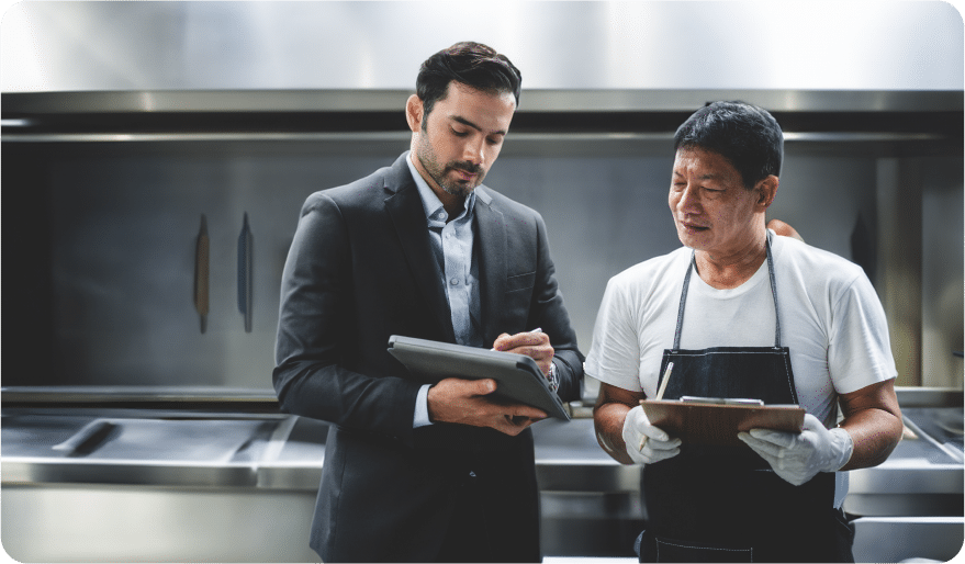 Un homme en costume montre quelque chose sur une tablette numérique à un homme en chemise blanche et tablier qui tient un bloc-notes ; ils se trouvent tous deux dans une cuisine professionnelle.