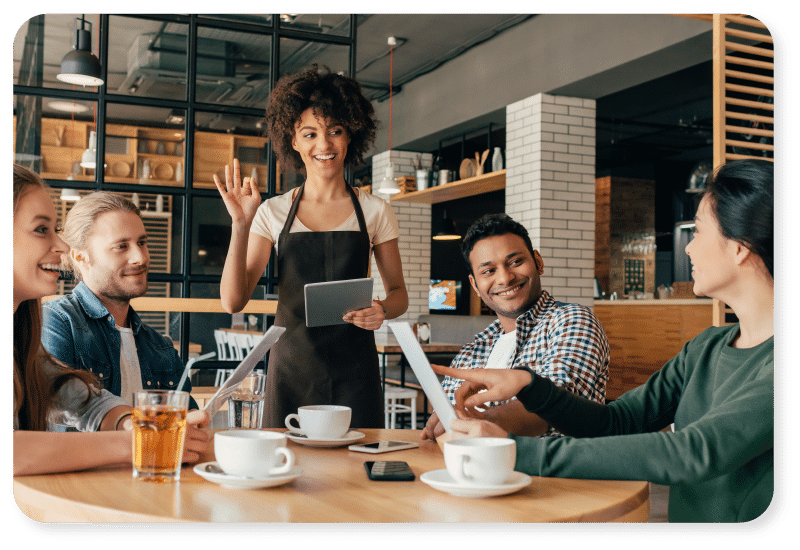 Un serveur souriant, muni d'un bloc-notes, vient accueillir quatre personnes assises à une table de café, devant leur café, leur thé et leurs menus. Le groupe semble heureux et à l'aise dans ce restaurant moderne et chaleureux.