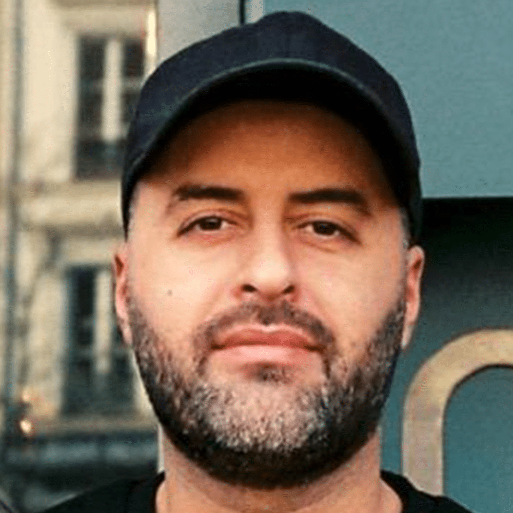Un homme à la barbe et à la moustache soigneusement taillées porte une casquette noire et une chemise foncée ; il se tient debout devant un immeuble doté de grandes fenêtres et d'un mur aux teintes verdâtres.