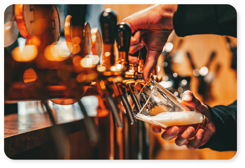 Dans un bar, une personne sert de la bière à partir d'un robinet dans un verre ; on aperçoit plusieurs robinets dorés et l'ambiance est chaleureuse et accueillante.