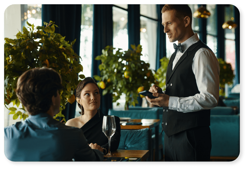 Un serveur vêtu d'un gilet noir prend la commande d'un couple assis dans un restaurant, avec des plantes vertes et de grandes baies vitrées en arrière-plan.