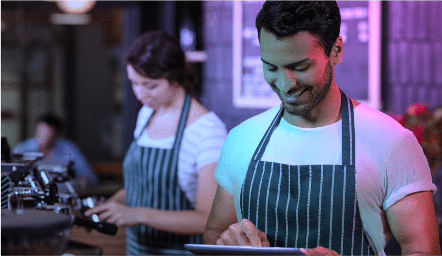 Un homme souriant, vêtu d'un tablier rayé, utilise une tablette, tandis qu'une femme portant un tablier similaire travaille à l'arrière-plan dans un café ou un salon de thé.