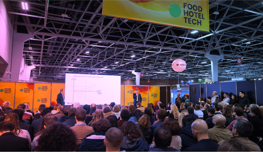 Un public nombreux assiste à une présentation au salon Food Hotel Tech, où deux intervenants se trouvent sur scène, devant un écran de projection et des bannières orange, dans un lieu spacieux et moderne.