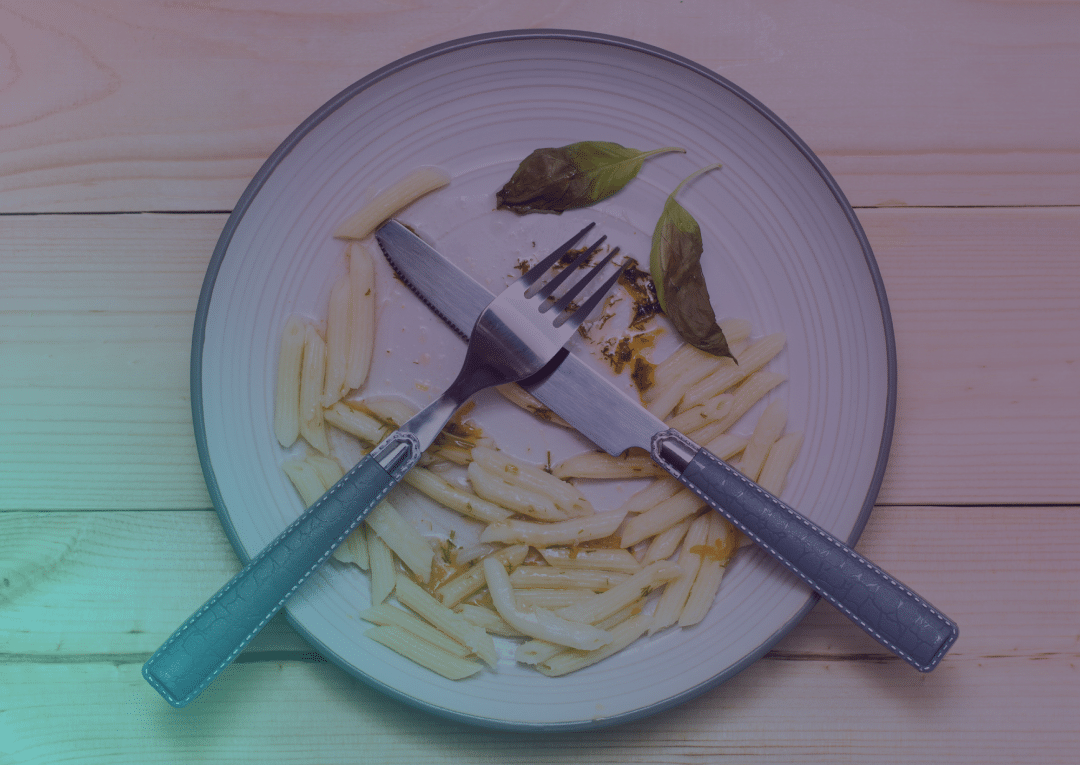 Une assiette presque vide, contenant quelques morceaux de penne, deux feuilles de basilic, ainsi qu'une fourchette et un couteau croisés au centre, posée sur une table en bois clair.