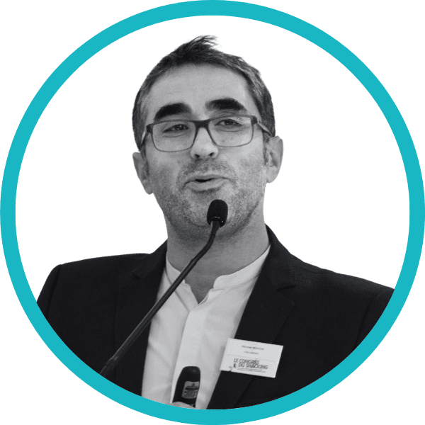 Un homme à lunettes, vêtu d'une veste de costume et d'une chemise blanche, parle dans un micro. Il porte un badge nominatif sur sa veste. L'image est en noir et blanc et entourée d'un cadre circulaire bleu sarcelle.