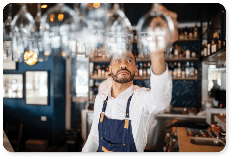 Un barman, vêtu d'une chemise blanche et d'un tablier bleu, tend le bras pour attraper un verre à vin suspendu derrière le comptoir, avec des étagères remplies de bouteilles visibles à l'arrière-plan.