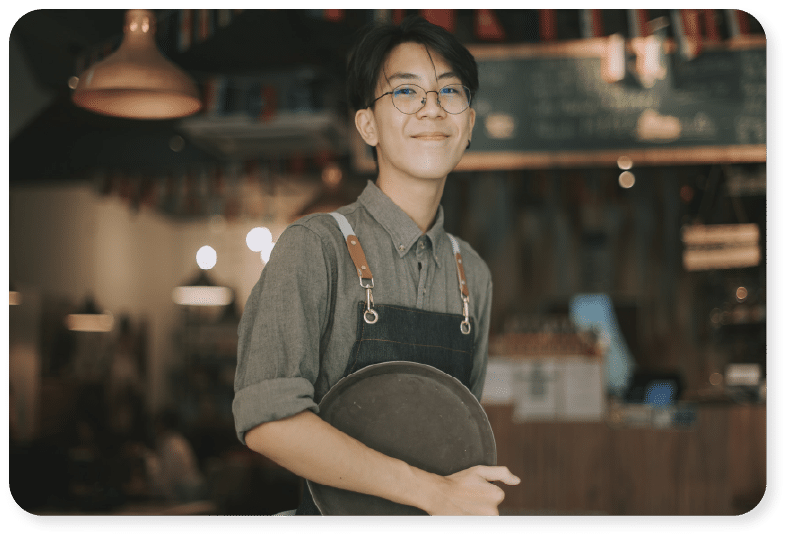 Une personne souriante, portant des lunettes, une chemise grise et un tablier foncé, tient un plateau rond dans un café à l'éclairage chaleureux, avec un arrière-plan flou.