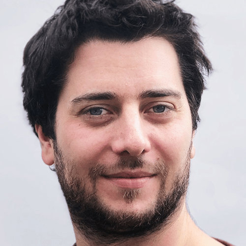 Un homme aux cheveux courts et foncés, à la peau claire et aux yeux clairs, sourit doucement à la caméra. L'arrière-plan est d'un gris clair uni.