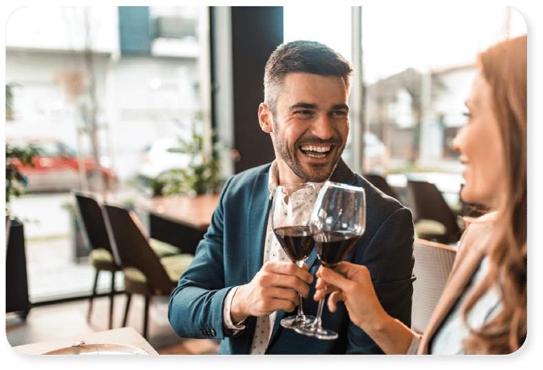 Un homme et une femme sont assis à une table de restaurant ; ils sourient et trinquent avec des verres de vin rouge. La lumière vive du jour pénètre par les grandes baies vitrées en arrière-plan.