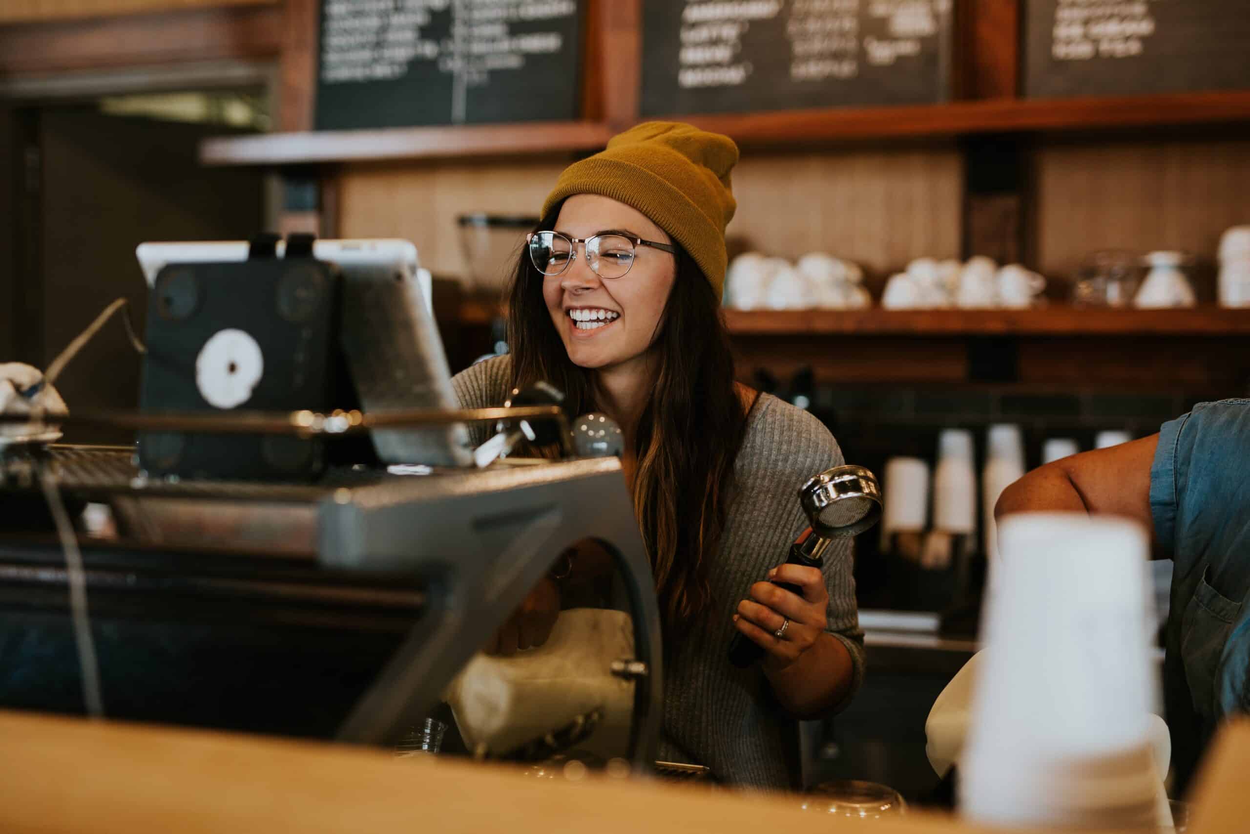 Un barista souriant, portant des lunettes et un bonnet jaune, se tient derrière le comptoir d'un café, un porte-filtre à la main, les yeux rivés sur la caisse. À l'arrière-plan, on aperçoit des étagères remplies de tasses et un panneau affichant le menu.