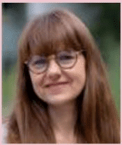 Une femme aux longs cheveux châtains et à la frange, portant des lunettes rondes, sourit doucement à la caméra. L'arrière-plan est constitué d'une végétation floue.