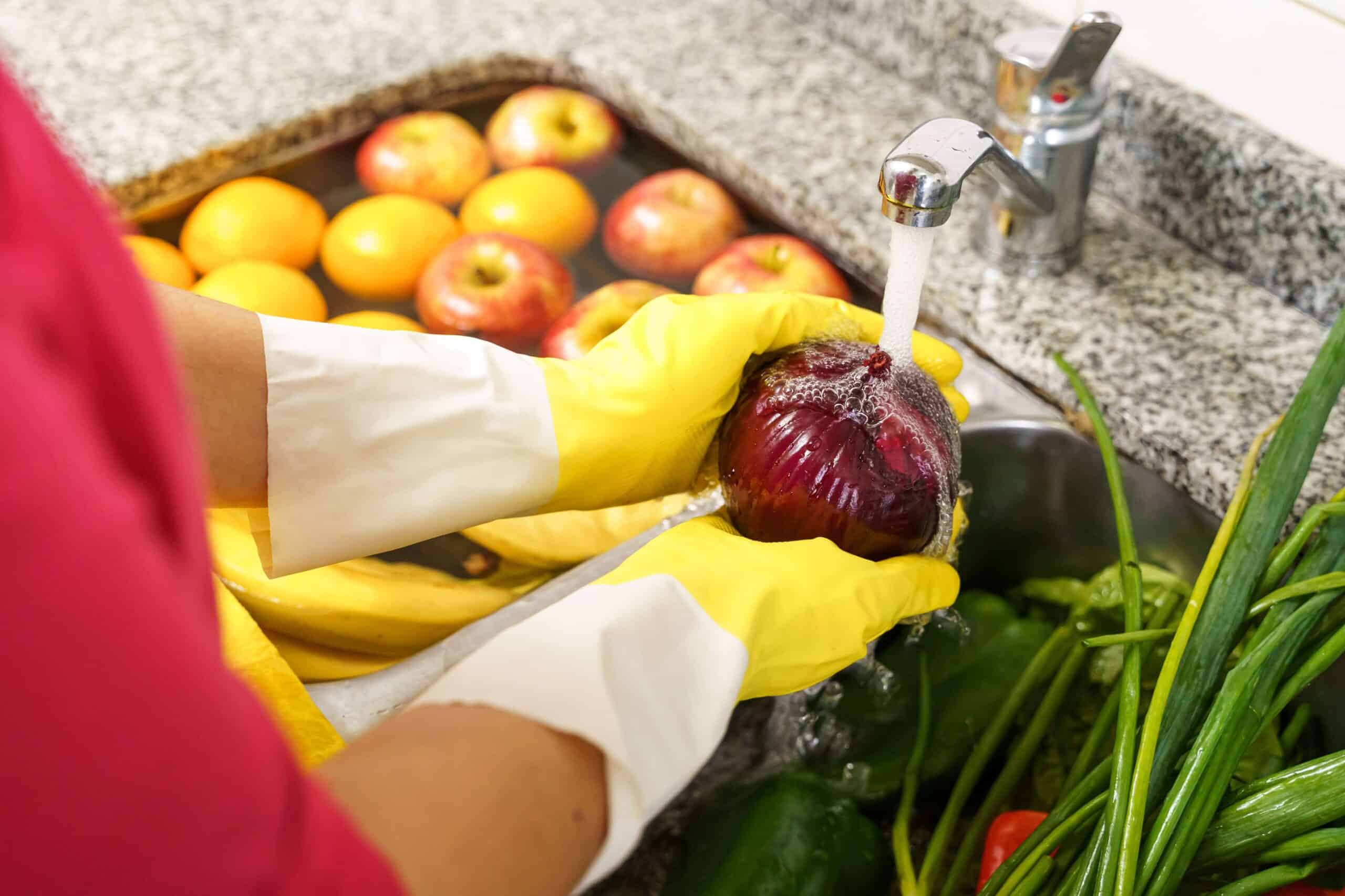 Une personne portant des gants jaunes rince un oignon rouge sous l'eau courante dans l'évier de la cuisine, à côté d'autres fruits et légumes frais.