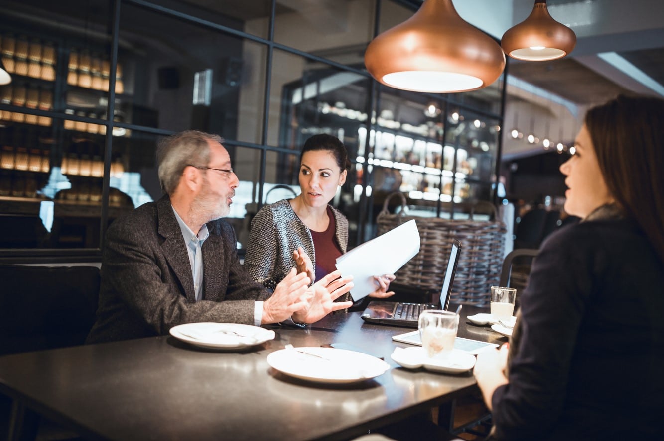 Trois personnes sont assises à une table de restaurant et discutent. Un homme gesticule en parlant, une femme tient des papiers et une autre femme écoute. Des assiettes et des verres sont posés sur la table.