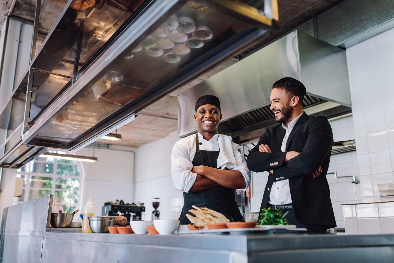 Deux hommes se tiennent debout, souriants, dans une cuisine professionnelle. L'un porte un uniforme de chef et un tablier, tandis que l'autre est en costume. Divers ingrédients alimentaires et ustensiles de cuisine sont visibles sur le plan de travail devant eux.