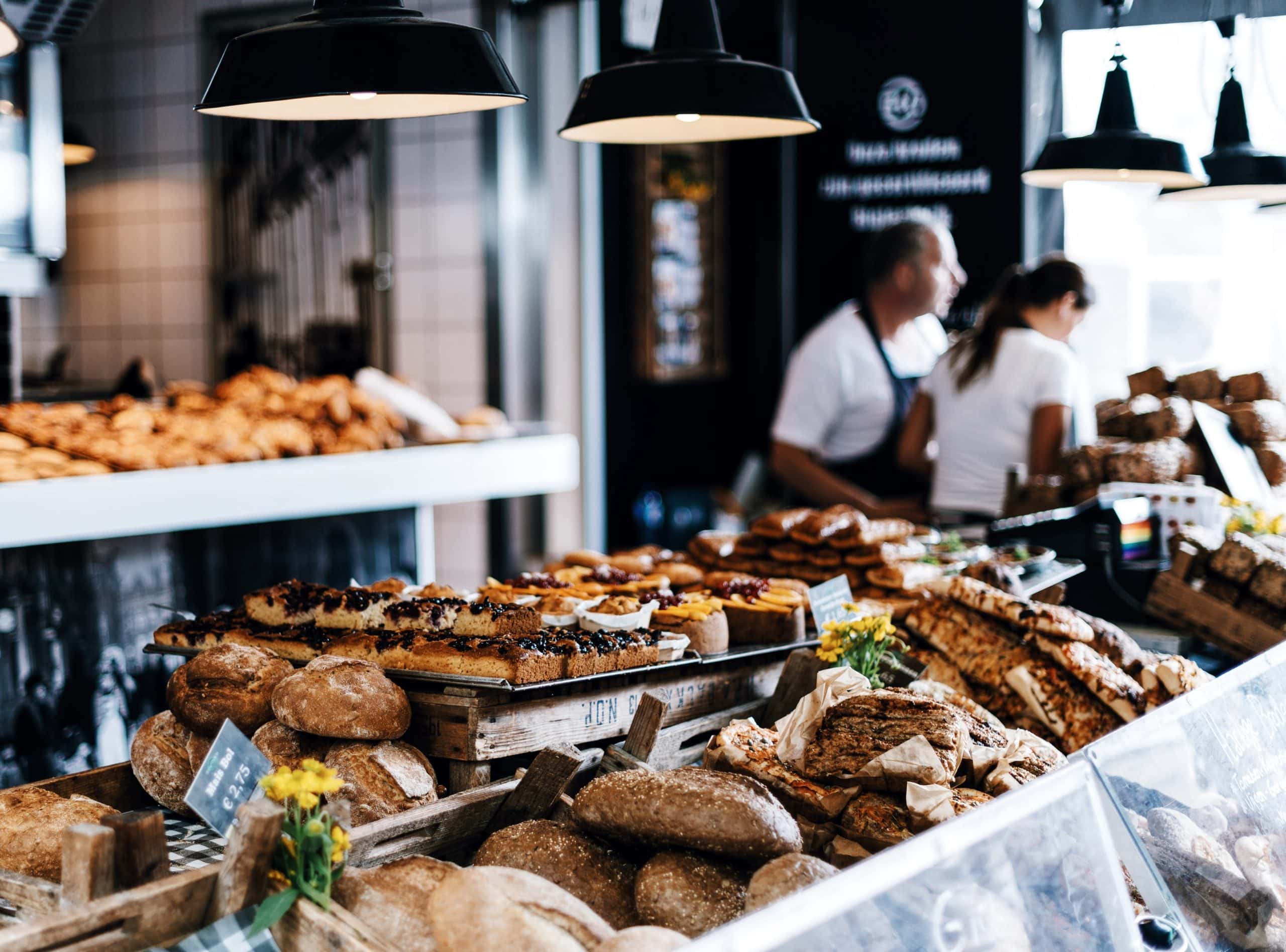 Une vitrine de boulangerie regorgeant de pains et de viennoiseries variés, avec deux personnes travaillant derrière le comptoir et des guirlandes lumineuses suspendues au-dessus.