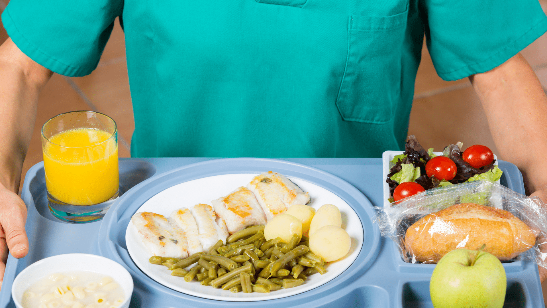 Une personne vêtue d'une blouse bleu sarcelle tient un plateau-repas composé de poisson, de pommes de terre, de haricots verts, de salade, de pain, d'une pomme, d'un verre de jus d'orange et d'un petit bol de pâtes.