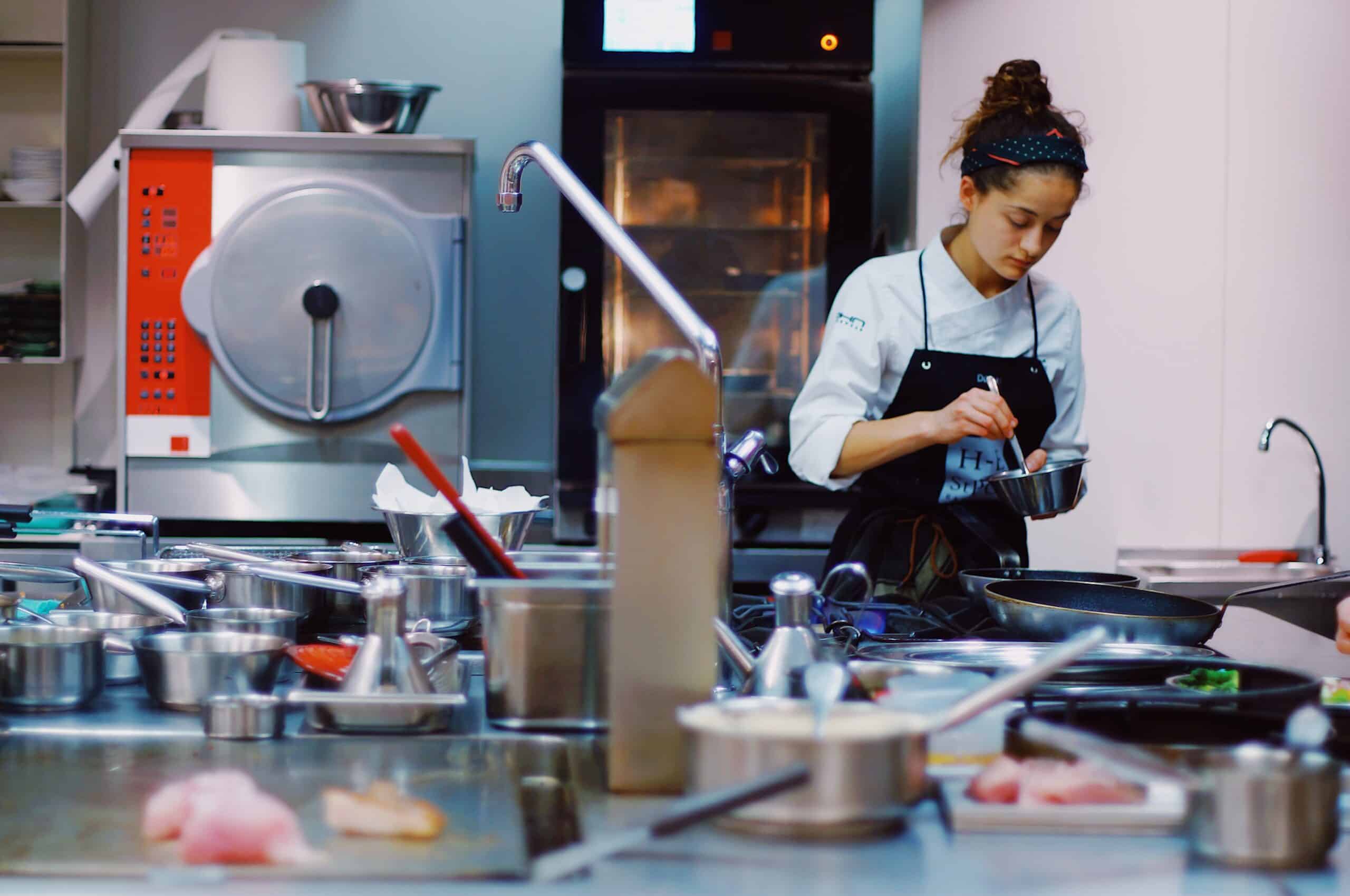 Un chef, vêtu d'une veste blanche et d'un tablier noir, prépare un plat devant un plan de travail en inox, entouré de divers ustensiles, bols et ingrédients. La cuisine semble propre et professionnelle.