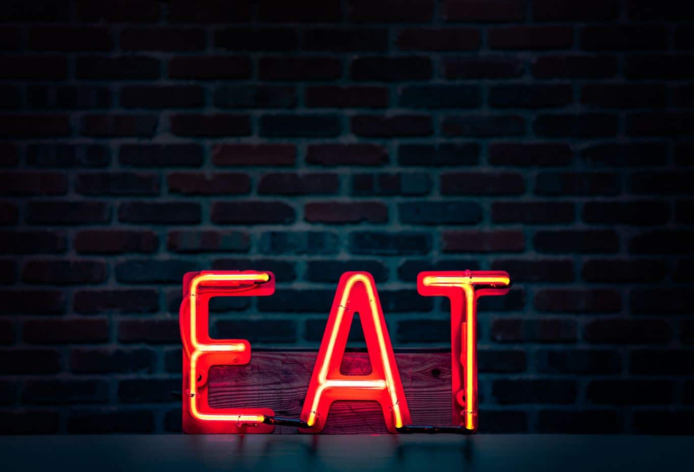 Une enseigne au néon rouge formant le mot « EAT » s'illumine sur fond de mur de briques sombres.