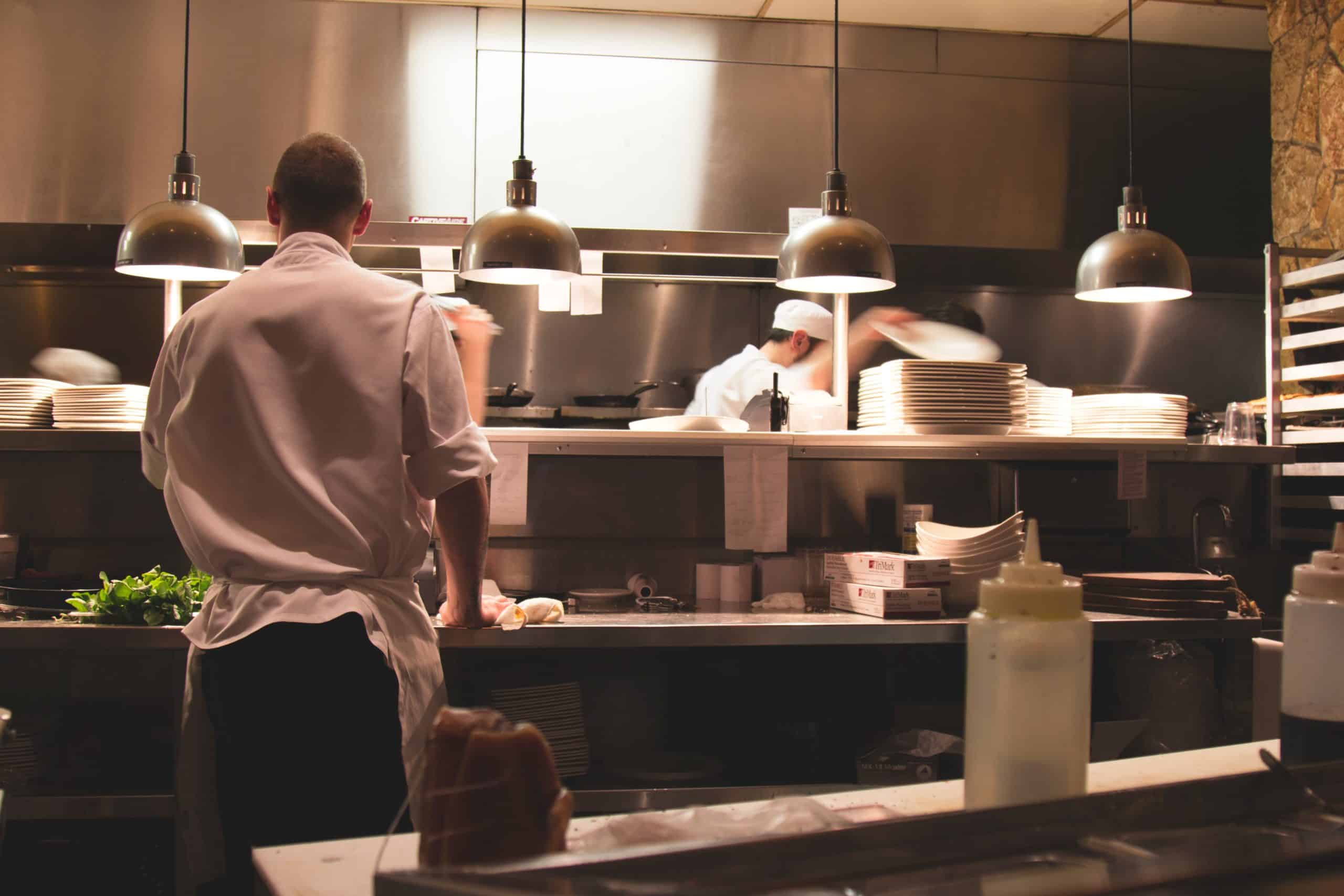 Un chef se tient debout devant un plan de travail de cuisine, de dos, tandis qu'à l'arrière-plan, d'autres membres du personnel s'affairent à empiler des assiettes sous des lampes suspendues qui diffusent une lumière chaleureuse.