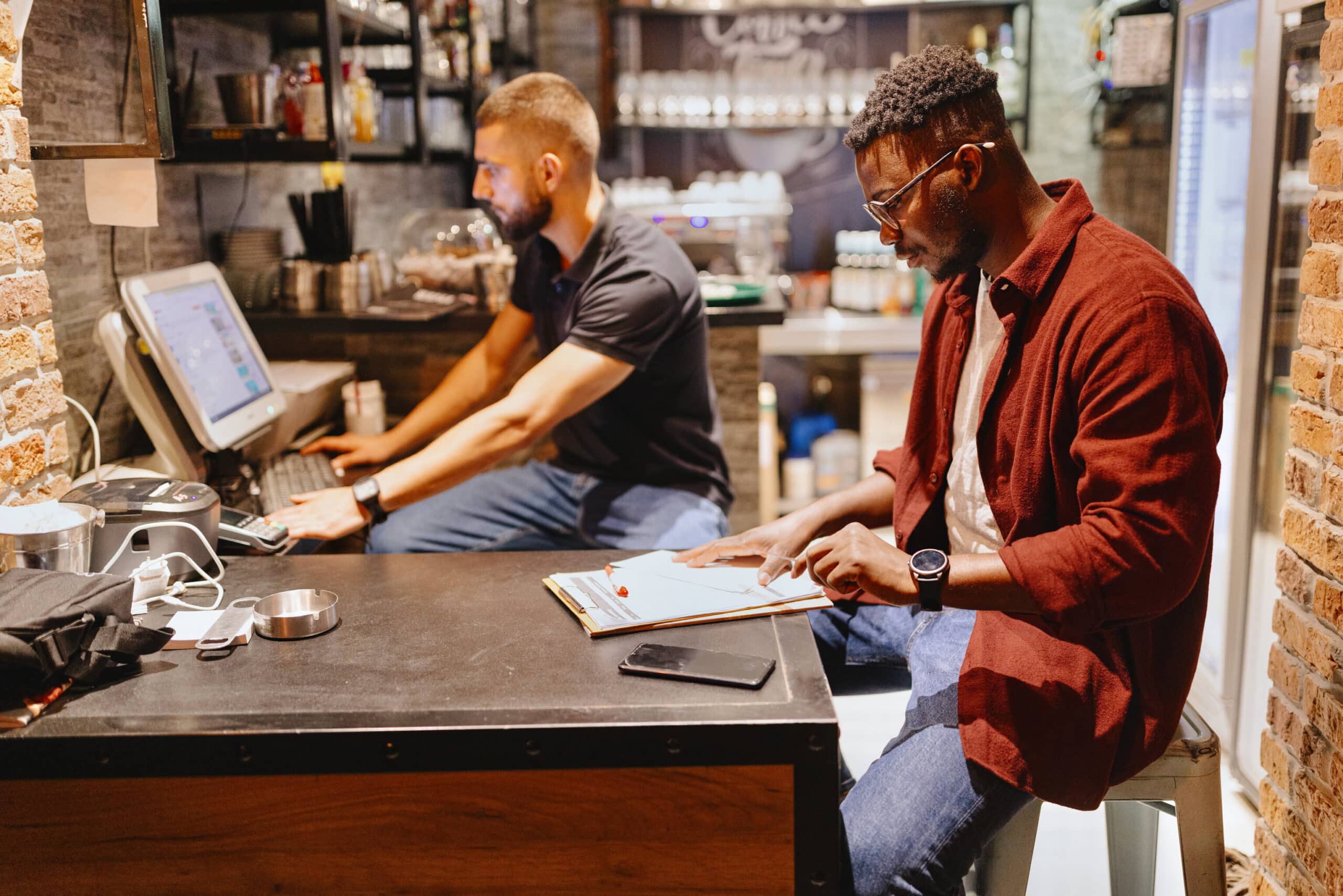Deux hommes travaillent dans un café ; l'un utilise un ordinateur au comptoir, l'autre prend des notes sur un bloc-notes. La pièce comporte des étagères, une machine à café et des murs en briques apparentes.