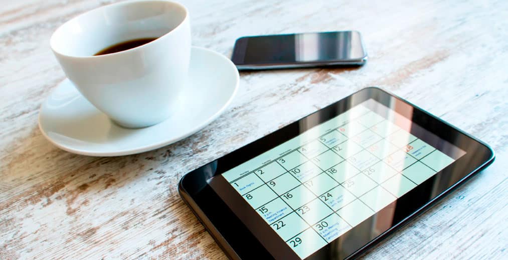 Sobre una mesa de madera, junto a una taza de café y un smartphone, hay una tableta en la que se ve un calendario.