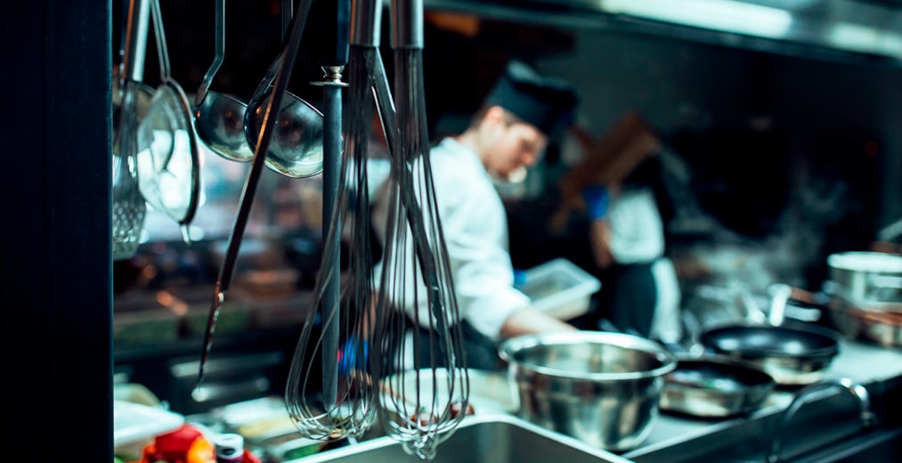 En primer plano cuelgan utensilios de cocina de metal, como batidores y cucharones. En el fondo, difuminado, un cocinero con uniforme blanco prepara la comida en una cocina moderna y ajetreada.