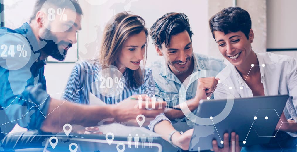 Cuatro personas sonrientes y trabajando en equipo alrededor de un ordenador portátil, con gráficos digitales de números, porcentajes e iconos superpuestos, lo que sugiere un análisis de datos o un trabajo en equipo en un entorno de oficina moderno.