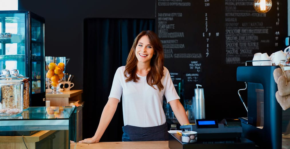 Una mujer está de pie, sonriendo, detrás del mostrador de una cafetería moderna, con una vitrina de pasteles y una pizarra con el menú al fondo.