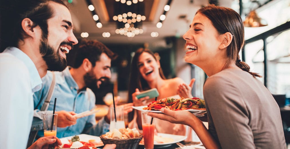 Cuatro amigos están sentados en la mesa de un restaurante, riendo y disfrutando juntos de la comida y la bebida. Una mujer sostiene un plato de pasta y sonríe radiante mientras el grupo comparte un momento de alegría.