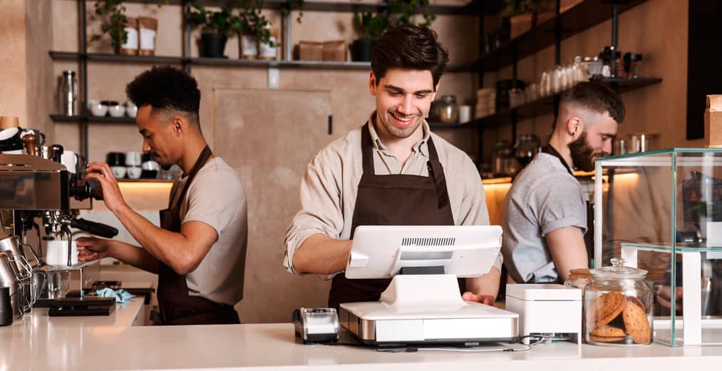 Tres baristas trabajando detrás del mostrador de una cafetería; uno sonríe junto a la caja registradora, otro maneja una máquina de café expreso y el tercero prepara algo en la trastienda.