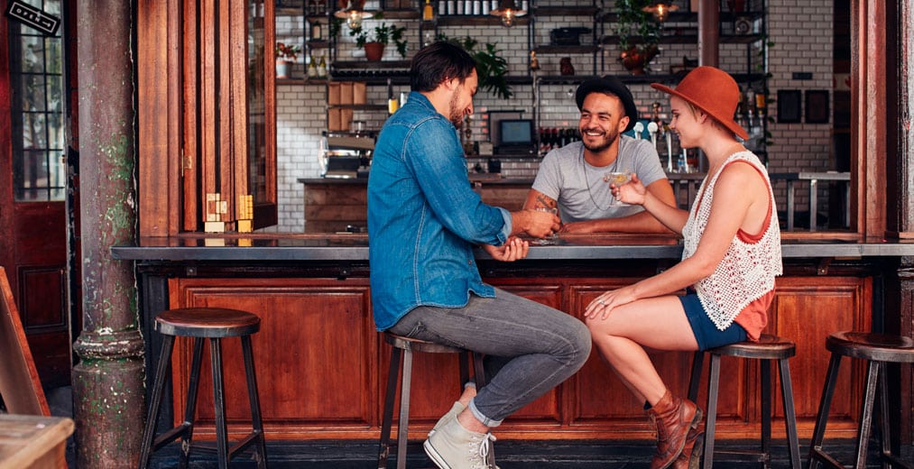 Tres personas están sentadas en la barra de un bar, sonriendo y charlando. Dos de ellas están sentadas en taburetes, una frente a la otra, mientras que el camarero está de pie detrás de la barra, hablando con ellas. El bar tiene paneles de madera y estanterías con botellas y plantas.