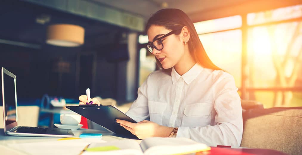 Una mujer con gafas escribe en un portapapeles junto a un escritorio en el que hay un ordenador portátil, unos papeles y una taza, en una oficina moderna iluminada por la luz del sol.