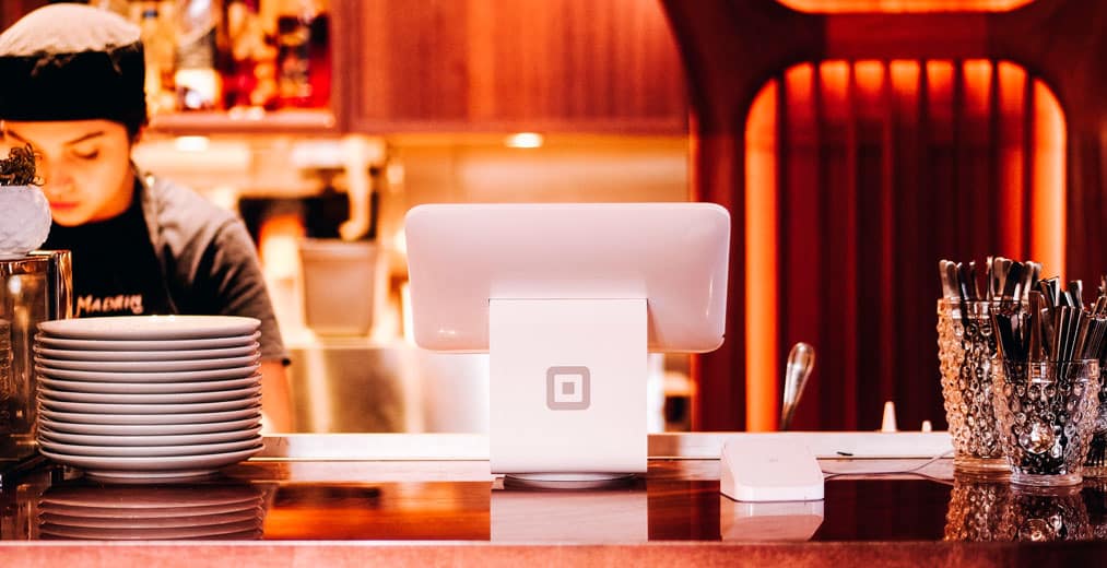 La barra de un restaurante con platos apilados, cubiertos en un soporte, un terminal de punto de venta Square y una persona trabajando al fondo. La iluminación cálida crea un ambiente acogedor.