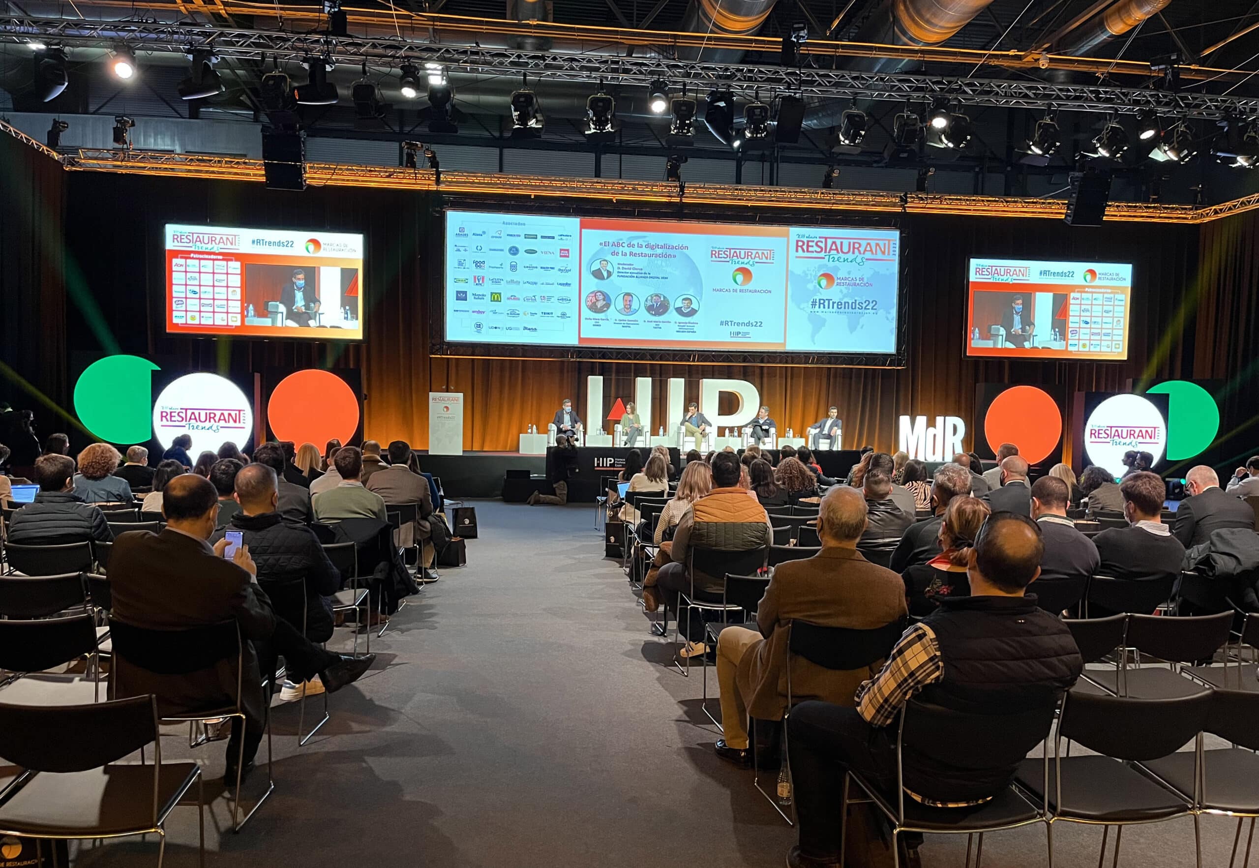 El público, sentado en una conferencia, asiste a una mesa redonda que tiene lugar en el escenario. En unas pantallas gigantes se muestra información sobre el evento. La sala está bien iluminada y cuenta con pancartas y elementos de imagen de marca relacionados con el evento.