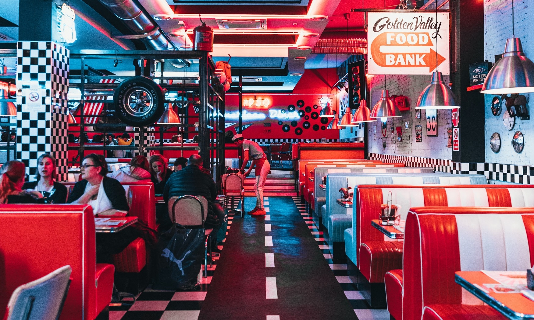 Una cafetería de estilo retro con reservados rojos y blancos, suelo a cuadros blancos y negros, luces de neón y un cartel del «Golden Valley Food Bank»; hay clientes sentados y una persona está limpiando cerca de la barra.