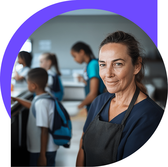 Una mujer sonriente, con un delantal oscuro, está de pie frente al mostrador de una cafetería, con unos niños y otro adulto al fondo esperando la comida.