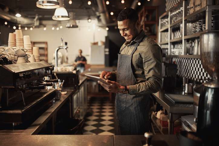 Un barista sonriente, que lleva un delantal, utiliza una tableta detrás de la barra de una cafetería moderna; al fondo se ven tazas, máquinas de café y estanterías.