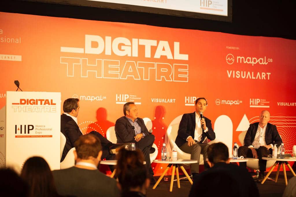 Cuatro hombres vestidos con traje están sentados en una mesa de ponentes sobre un escenario, frente a un telón de fondo naranja con la inscripción «DIGITAL THEATRE», dirigiéndose al público en un evento profesional. Se ven un atril y unas botellas de agua.