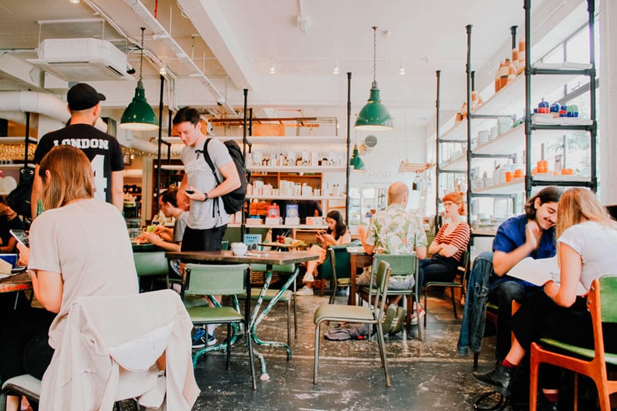 Una escena animada en una cafetería, con gente sentada en las mesas, charlando, utilizando ordenadores portátiles y tomando café. La iluminación brillante, las sillas verdes y las estanterías con libros y productos al fondo crean un ambiente animado.